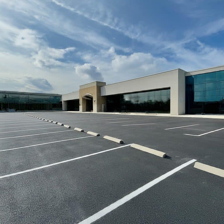 A large, empty parking lot with white parking lines in front of a modern-commercial building under a blue sky with scattered clouds.