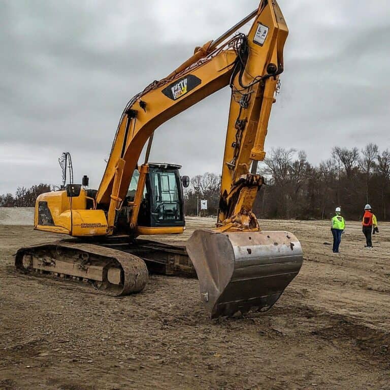 A yellow excavator is parked on a construction site with two workers wearing safety gear walking nearby.