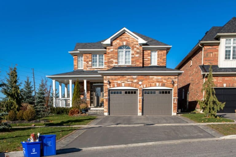 A two-story brick house with a double garage, driveway, and a front porch is seen on a clear day. A blue recycling bin and a brown paper bag are placed at the end of the driveway.
