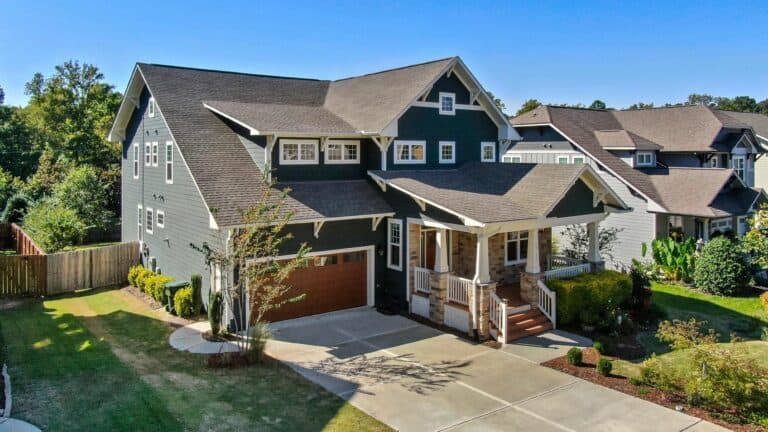 Two-story house with a grey exterior, a sloped roof, and a wooden front porch. It features a large driveway leading to an attached garage and is surrounded by a well-maintained yard and shrubs.