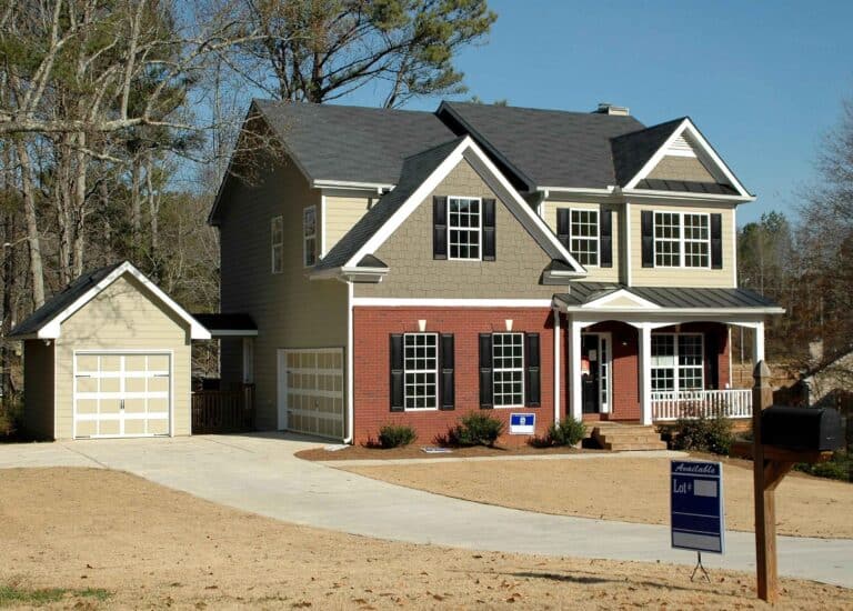 A two-story house with a mix of brick and siding exterior, a driveway leading to a garage, and a "For Sale" sign in the front yard. The property includes a lawn with some trees in the background.