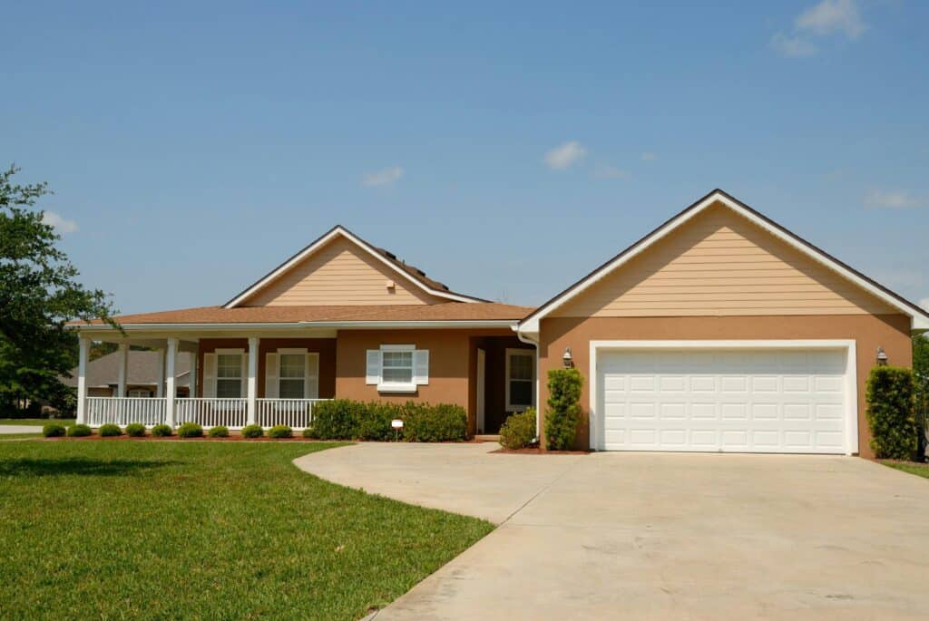 Single-story house with beige siding, a white front porch, and a two-car garage. A well-maintained lawn and concrete driveway are in the foreground, with a bright blue sky above.