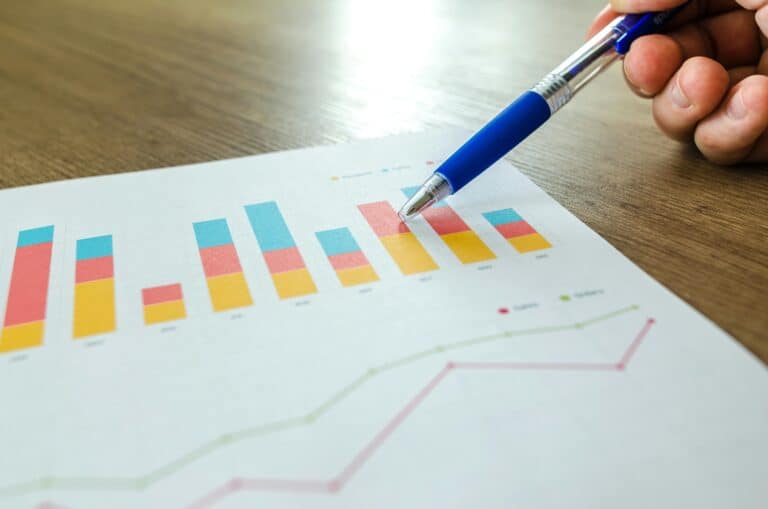 A hand holding a pen points to a bar graph on a piece of paper on a wooden table, with another line graph visible underneath.