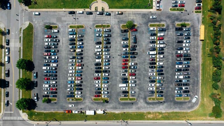 Aerial view of a large parking lot filled with rows of parked cars and a yellow school bus near the upper right corner, surrounded by green grass and trees.