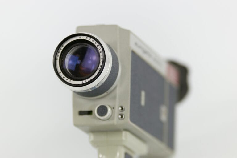 Close-up of a vintage video camera lens with a metallic body. The lens shows the brand "Angenieux" and specifications. The background is blurred.